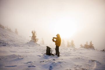 Hiker in yellow jacket opening thermos with hot drink at snowy mountain during misty sunset. Refreshment while resting on winter hike
