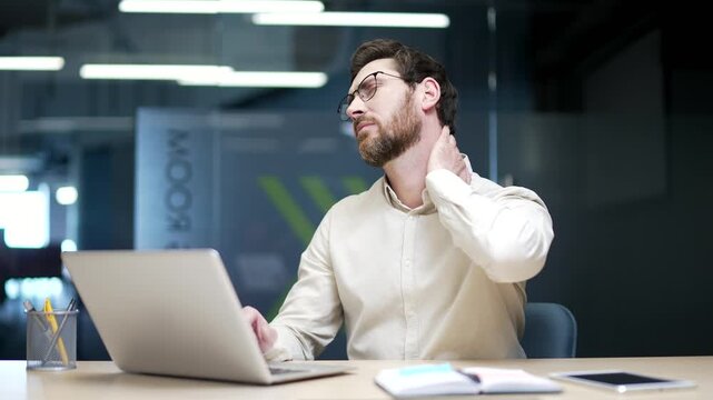 Tired businessman sitting at desk in modern office working on laptop and suffering from neck pain. Upset male employee feeling discomfort, massaging sore muscles from stress, tension, or poor posture.