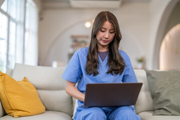 Asian nurse wearing blue scrubs using laptop computer on sofa, Young female doctor working online or studying, Telemedicine, digital healthcare, medical education and remote work concept