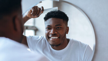 A man smiles while using a comb to style his hair in a bathroom. The scene shows him in a white shirt and the light coming from a nearby window.