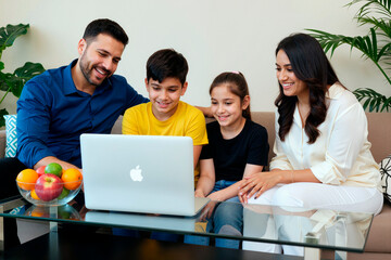 Fototapeta premium Hispanic man, woman, boy and girl sitting together on sofa smiling and watching laptop screen, family spending time together using computer in modern living room