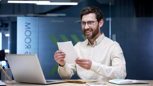 Smiling surprised businessman reading letter with great news sitting at desk in office. Excited joyful employee celebrating success, feeling happiness and achievement after receiving positive message