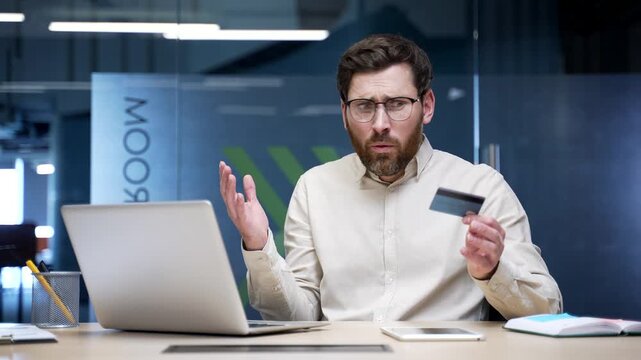Shocked upset businessman sitting at desk in modern office checking credit card on laptop. Frustrated male discovering fraud or online scam, feeling angry and disappointed after financial deception