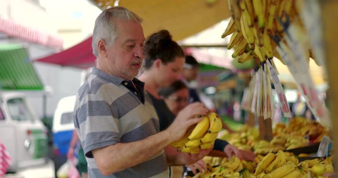 Elderly man selecting bananas at outdoor street market carefully choosing fruit during everyday local food shopping routine