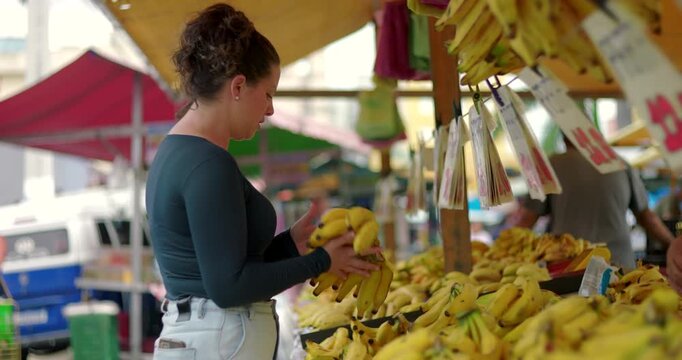 Young woman selecting bananas at outdoor street market holding fruit bunch and engaging with local produce display during everyday food shopping
