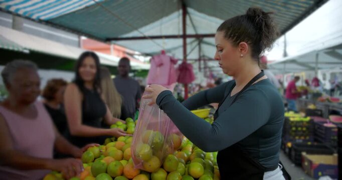 Female street vendor placing citrus fruit into plastic bag at outdoor market while customers wait conveying routine service and everyday local food commerce