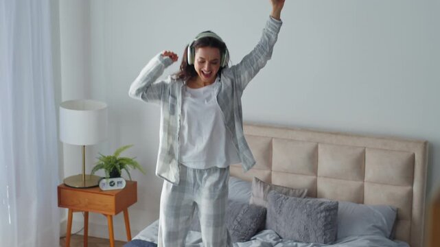 Carefree Caucasian woman jumping dancing on bed listening music in earphones smiling moving hands party jump in bedroom home female girl listen headphones audio song sound holding mobile phone delight