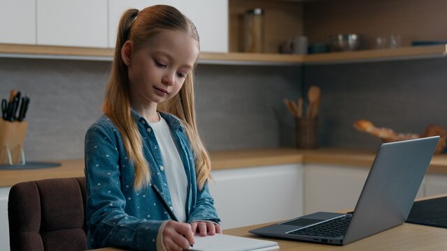 Caucasian kid girl school pupil e-learning in apartment homeschooling studying online in kitchen watching laptop class lesson course by video call writing homework child remote education quarantine