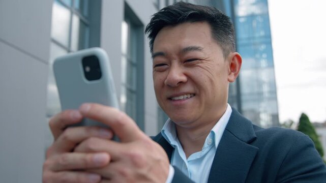 Positive Asian Korean business man Chinese middle-aged businessman with phone smiling laughing satisfied chatting smartphone in city downtown adult Japanese employer answer good mobile email online