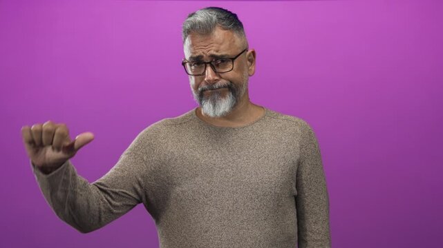 Man with gray beard and glasses points thumb to side with bare hand visible in studio against purple backdrop; skepticism self reflection.