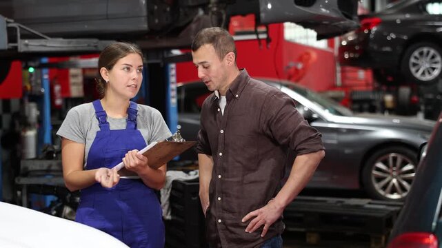 Positive young girl in blue overalls holding sheets and man client of car workshop communicating about service fees