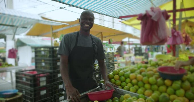 African American street vendor leaning on produce stall smiling at camera reflecting ease pride and belonging within neighborhood street market environment