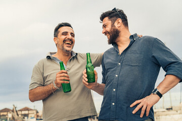 Two adult men laughing and drinking beer together outdoors