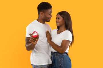 A couple stands close together, smiling and looking at each other. One partner holds a ring box while the other gently touches their partner's arm. They stand against a bright yellow background.