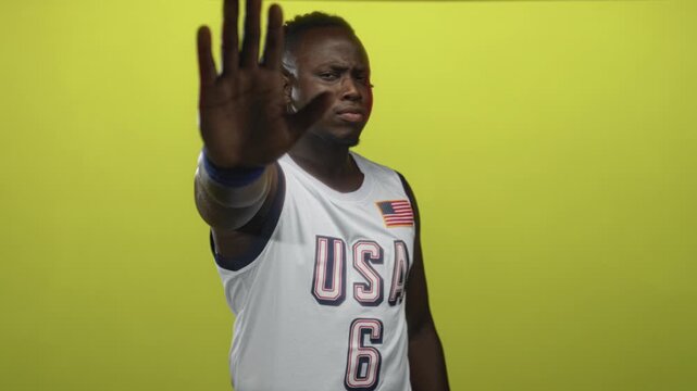 Man with usa basketball jersey showing palm to camera stop gesture in studio; defiance assertiveness boundary.