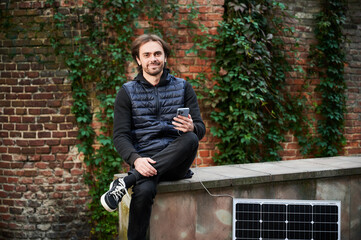 Handsome man using smartphone connected to photovoltaic solar panel. Integration of sustainable renewable energy into everyday life, demonstrating practical use of solar power for charging devices.