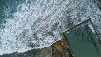 Aerial view of the turquoise ocean pool meets the wild, foamy waves crashing against the shore, creating a stark contrast in color and texture, Sydney, New South Wales, Australia.