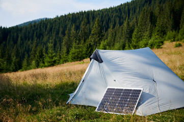 White tent equipped with solar panel sits in grassy field surrounded by lush trees and hills. Solar panel highlights eco-friendly approach to camping in tranquil, serene natural setting.
