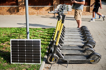 Row of electric scooters parked beside solar panel in outdoor urban area. Integration of solar power as sustainable energy source for charging electric scooters, promoting eco-friendly transportation.