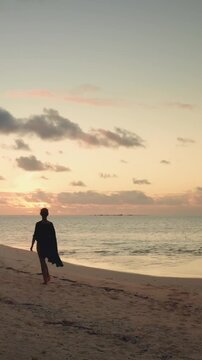 Silhouette of a person walking along a serene tropical beach during sunset, with palm trees framing the scene and a boat anchored near the shore. Peaceful ocean and island in the distance