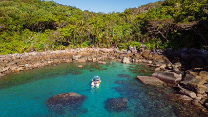 Aerial view of a boat floating on the crystalline turquoise waters near a rocky shore covered with lush, green tropical forest, Mission Beach, Queensland, Australia.
