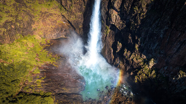 Aerial view of Wallaman Falls plunging into a turquoise pool, framed by rugged cliffs and lush vegetation, a vibrant rainbow arcing at its base, Wallaman, Queensland, Australia.