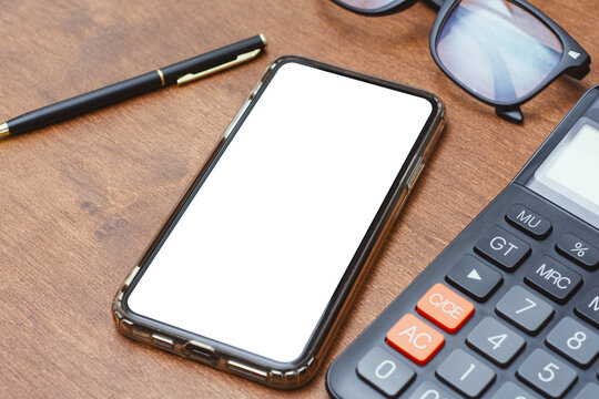Mobile phone mockup with an empty white screen on a wooden office table next to a calculator and stationery. Minimalist scene for app interfaces, digital banking and workspace branding.