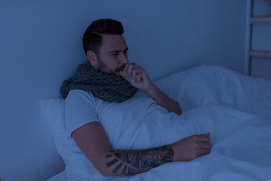 Coronavirus, influenza concept. Sick young man coughing, holding paper napkin near mouth, sitting in bed at night, feeling unwell, copy space