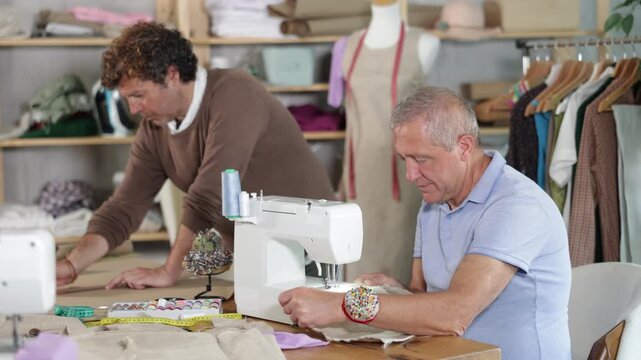 Elderly man tailor sews on machine adult man assistant drawing pattern on paper in workshop