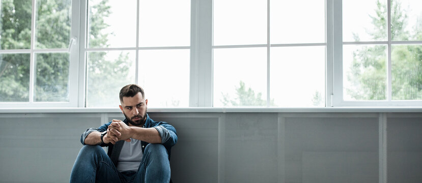 Man feeling sad, tired and worried, suffering depression in mental health, problems and broken heart. Frustrated young handsome bearded guy sitting on floor near large window, copy space, panorama