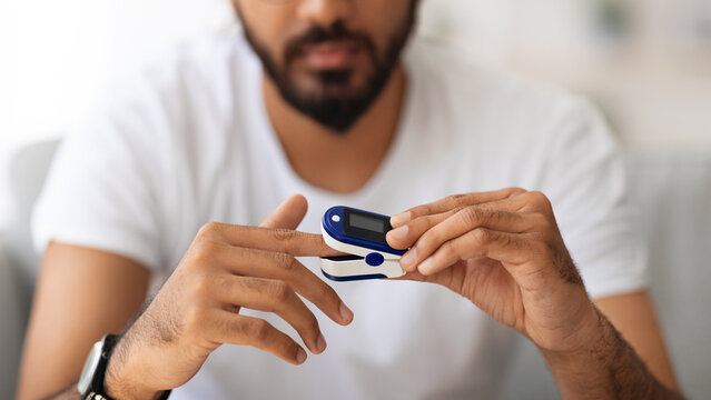 Closeup of unrecognizable man in white t-shirt using modern pulse oximeter at home, copy space. Cropped of middle-eastern male measuring his saturation, making self checkup with clip-like device