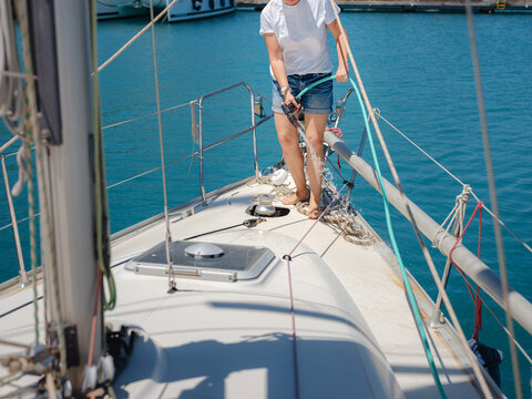 Adult woman washes teak deck of sailing yacht using water hose. Maintenance routine in Finike marina ensures boat remains clean during sunny summer day in Mediterranean Turkey.