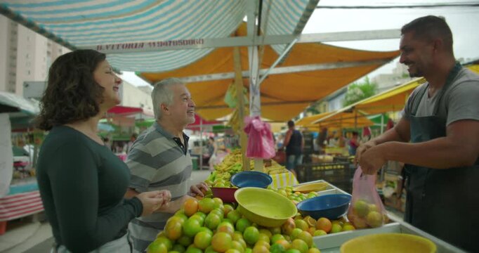 Young woman and elderly man conversing at open air market produce stall during routine food shopping reflecting everyday local commerce social exchange and familiar community rhythm