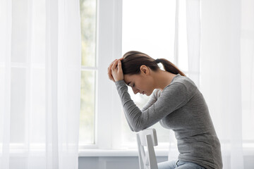 Unhappy european millennial female sad at home alone, holding her head and sitting on chair, on...