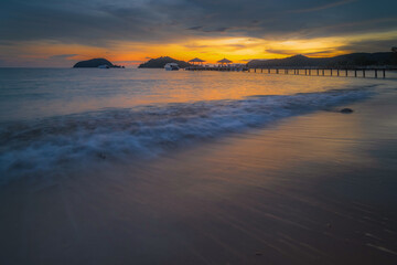 Fototapeta premium A beach with calm sea waves in the evening, a bridge in the background, and a sky with the sun setting.