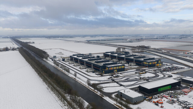 Aerial view of modern buildings contrasting against the snowy landscape, with long roads and a canal cutting through the scenery, Datacenter, Noord-Holland, Netherlands.