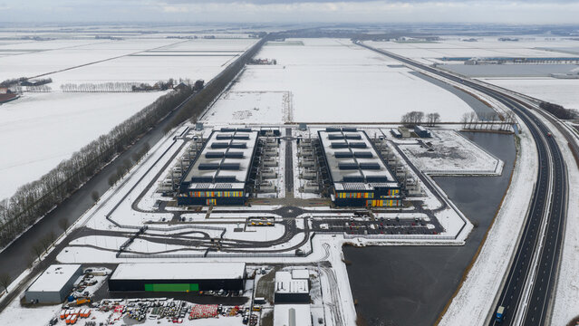 Aerial view of a modern datacenter complex amidst a stark, snow-covered landscape, contrasting with the dark asphalt of the adjacent highway, Datacenter, Noord-Holland, Netherlands.