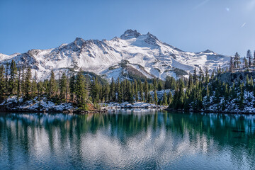 A mountain range is reflected in a lake. The sky is clear and blue. The scene is peaceful and serene