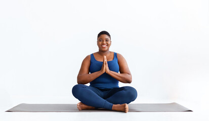 Fototapeta premium A person sits cross-legged on a mat with hands together in front of their chest. The setting is simple and bright with light walls. Focus is on yoga practice during the day.