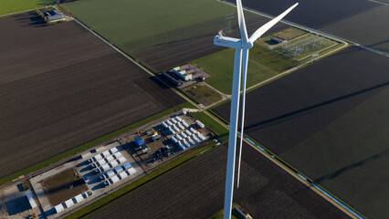 Dronten, Netherlands - 14 January 2026: Aerial view of a towering wind turbine casting a long shadow over the dark fields, adjacent to an energy storage facility filled with rows of white containers.