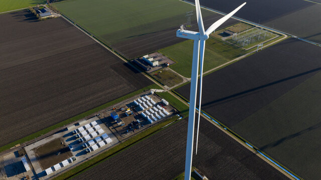Dronten, Netherlands - 14 January 2026: Aerial view of a towering wind turbine casting a long shadow over the dark fields, adjacent to an energy storage facility filled with rows of white containers.