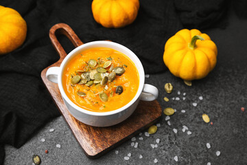 Warm pumpkin soup in a white bowl garnished with seeds on a wooden board with small pumpkins and dark cloth.