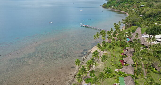 Savusavu resort with traditional bures huts and lush tropical landscape reef coastline. Exotic relaxing travel vacations and scenic views of the tranquil bay with boats and a long pier. Aerial flight