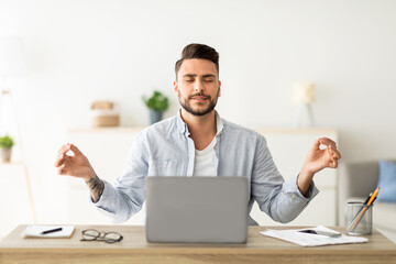 Keep calm. Male freelancer meditating while working on laptop computer, sitting with eyes closed, relaxing for stress relief at his workplace. Meditation and relaxation exercise concept