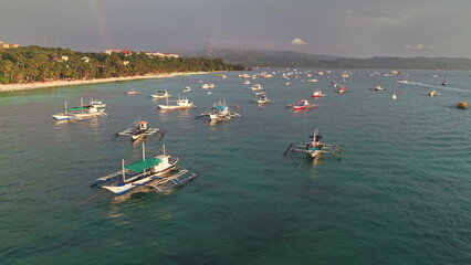 Aerial view of many colorful outrigger boats gently floating on turquoise water near a tropical beach on Boracay Island, Philippines, with a dramatic sky and a rainbow