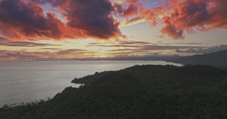 Lush tropical forest covers the hilly coastline of Savusavu, Fiji, creating a dramatic landscape under a vibrant sunset sky with warm orange and purple clouds reflecting over the calm Pacific Ocean