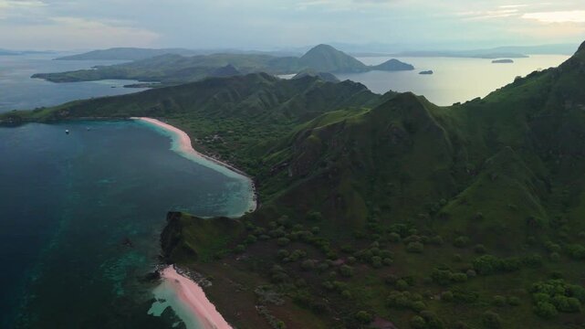 Drone footage of Padar Island in Komodo National Park, Indonesia, revealing the curved shoreline of Pink Beach, lush green hills and calm tropical sea during early morning light in 4K.