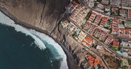 Tenerife: Los Gigantes dramatic rocky coastline town, Canary Islands, dark blue ocean waves breaking on the volcanic mountain hill shore. Resort hotel residential buildings top down, aerial flight