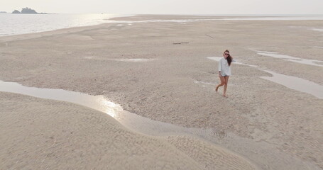 Woman enjoying leisurely barefoot walk on vast sandy beach at low tide, explore receding waters and textured patterns under the warm sunlight during a relaxing ocean vacation. Aerial drone flight
