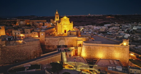 Malta, Comino island: Majestically illuminated historic Citadel in Gozo. The ancient fortifications glow beautifully in the night electric lights. Aerial view drone flight rotation panorama